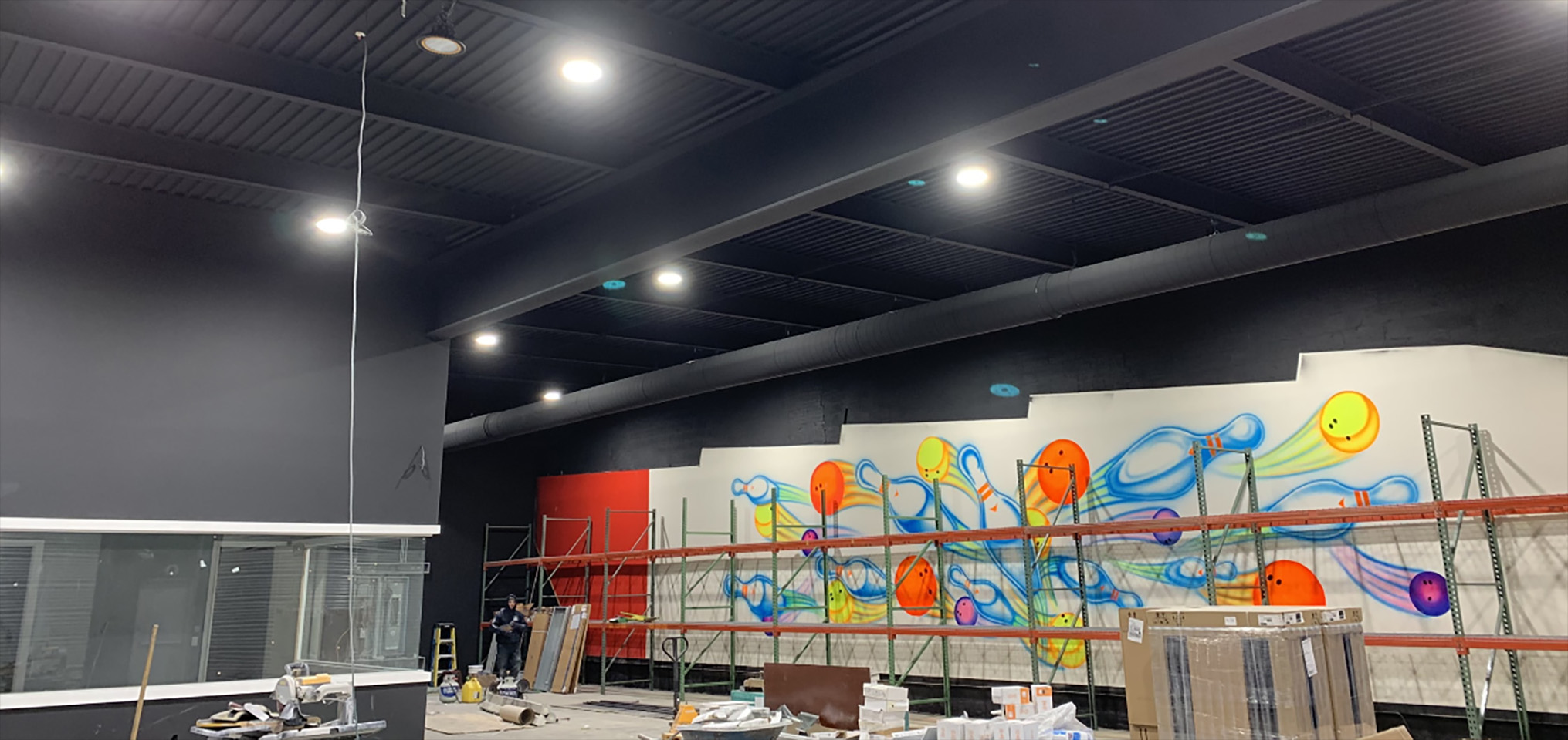 Commercial interior construction project with high ceilings, recessed lighting, shelving installation, and colorful wall mural artwork in progress.
